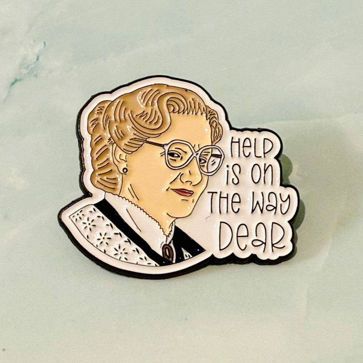 Enamel pin with a character wearing glasses and text 'Help is on the way dear' on a light background