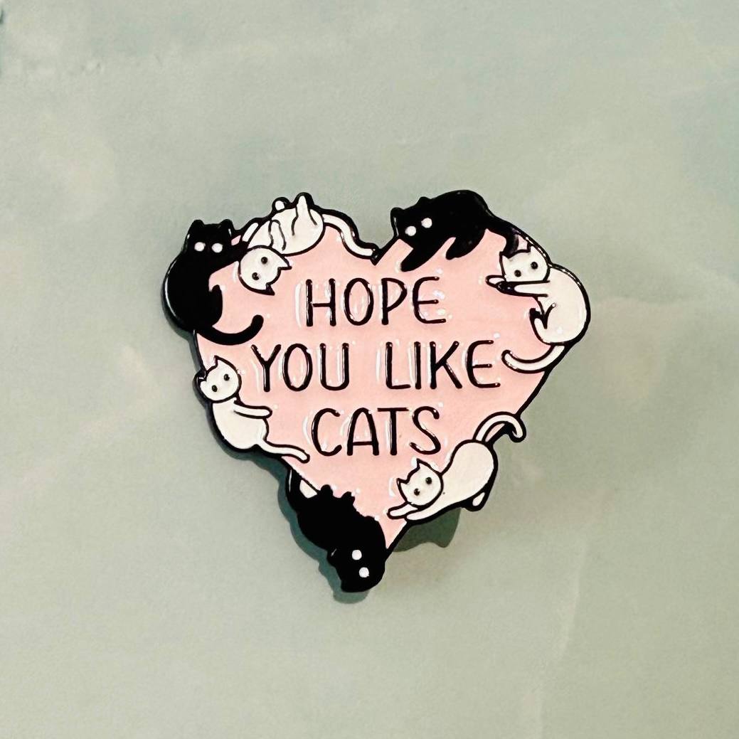 Heart-shaped pin with cartoon cats and 'Hope You Like Cats' text on a light gray background
