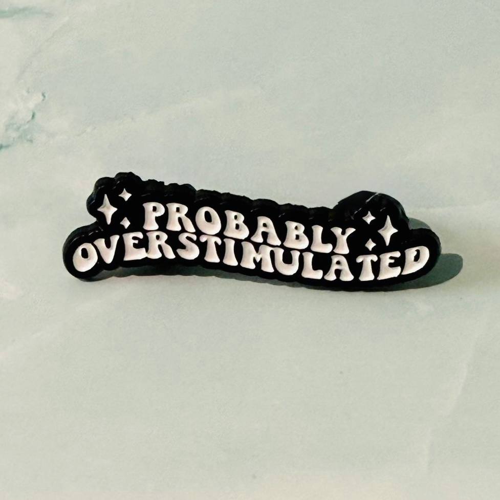 Black and white enamel pin with 'PROBABLY OVERSTIMULATED' text on a light gray background