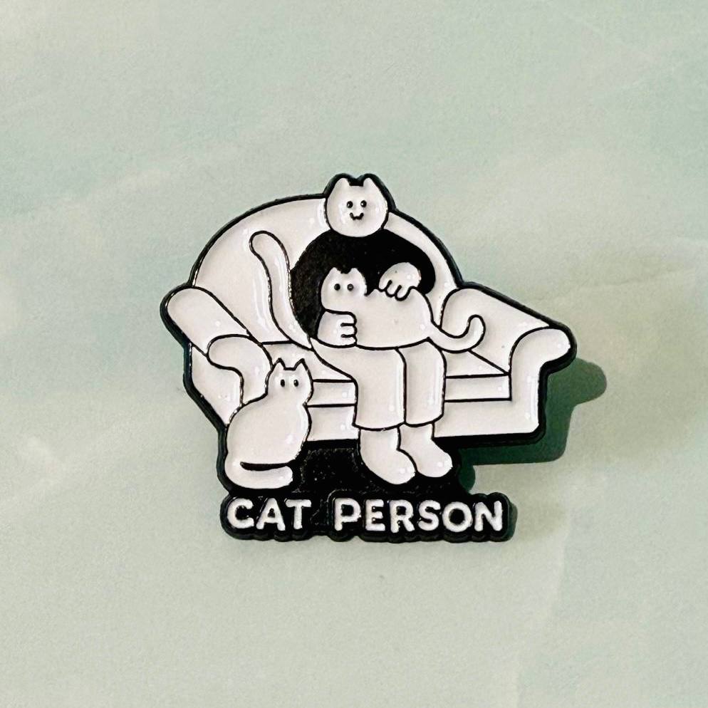 Pin with cartoon character sitting on a couch with a cat, labeled 'Cat Person' on a light gray background.
