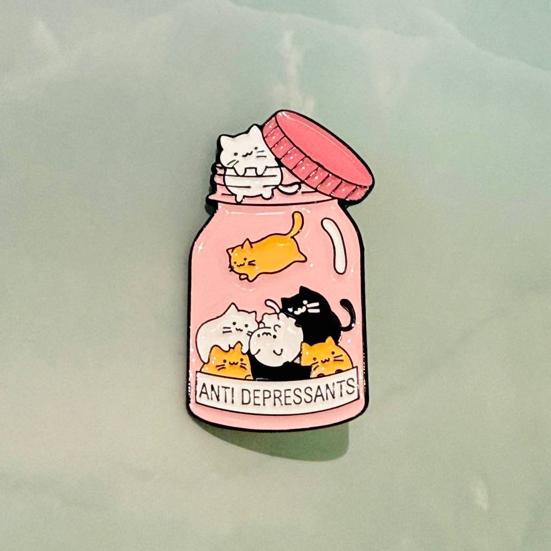 Pin with cartoon cats on a pink 'Anti Depressants' jar against a light green background