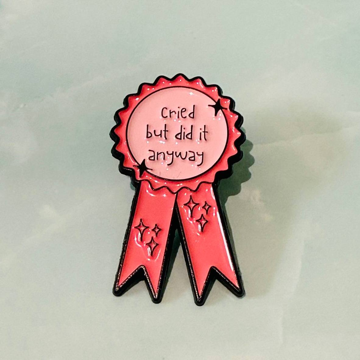 Pin with pink ribbon and text 'cried but did it anyway' on a light gray background