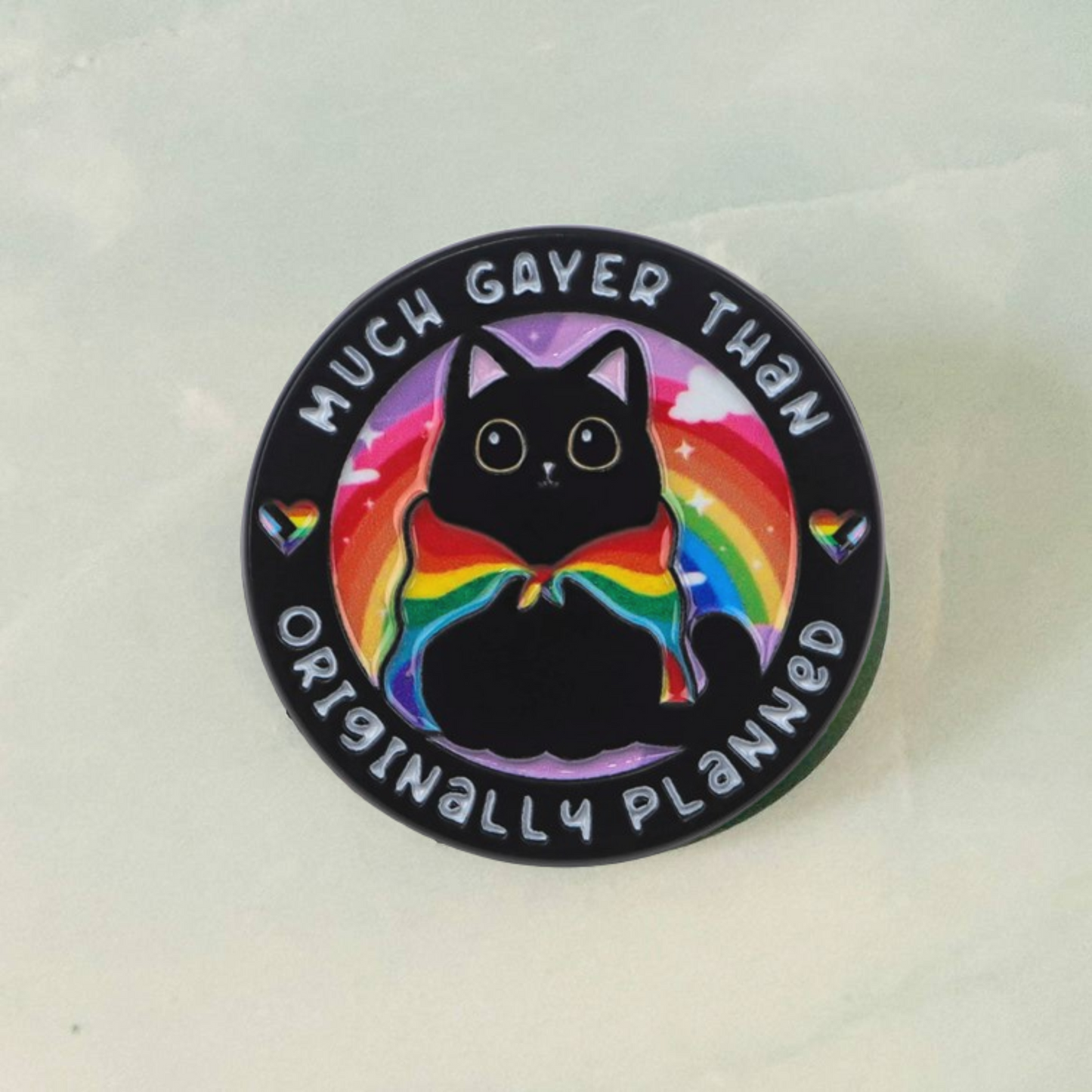 Button badge with a black cat wearing a rainbow cape and text 'Much Gayer Than Originally Planned' on a light background.