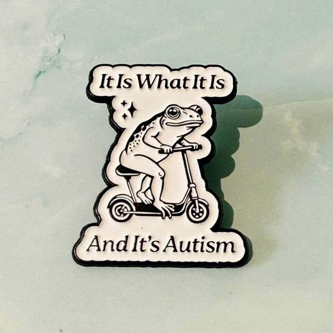 Pin with a frog on a scooter and text 'It Is What It Is And It's Autism' on a marble background