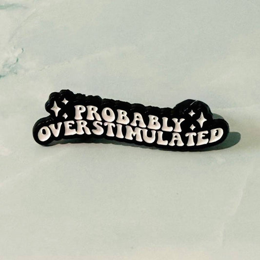 Black and white enamel pin with 'PROBABLY OVERSTIMULATED' text on a light gray background