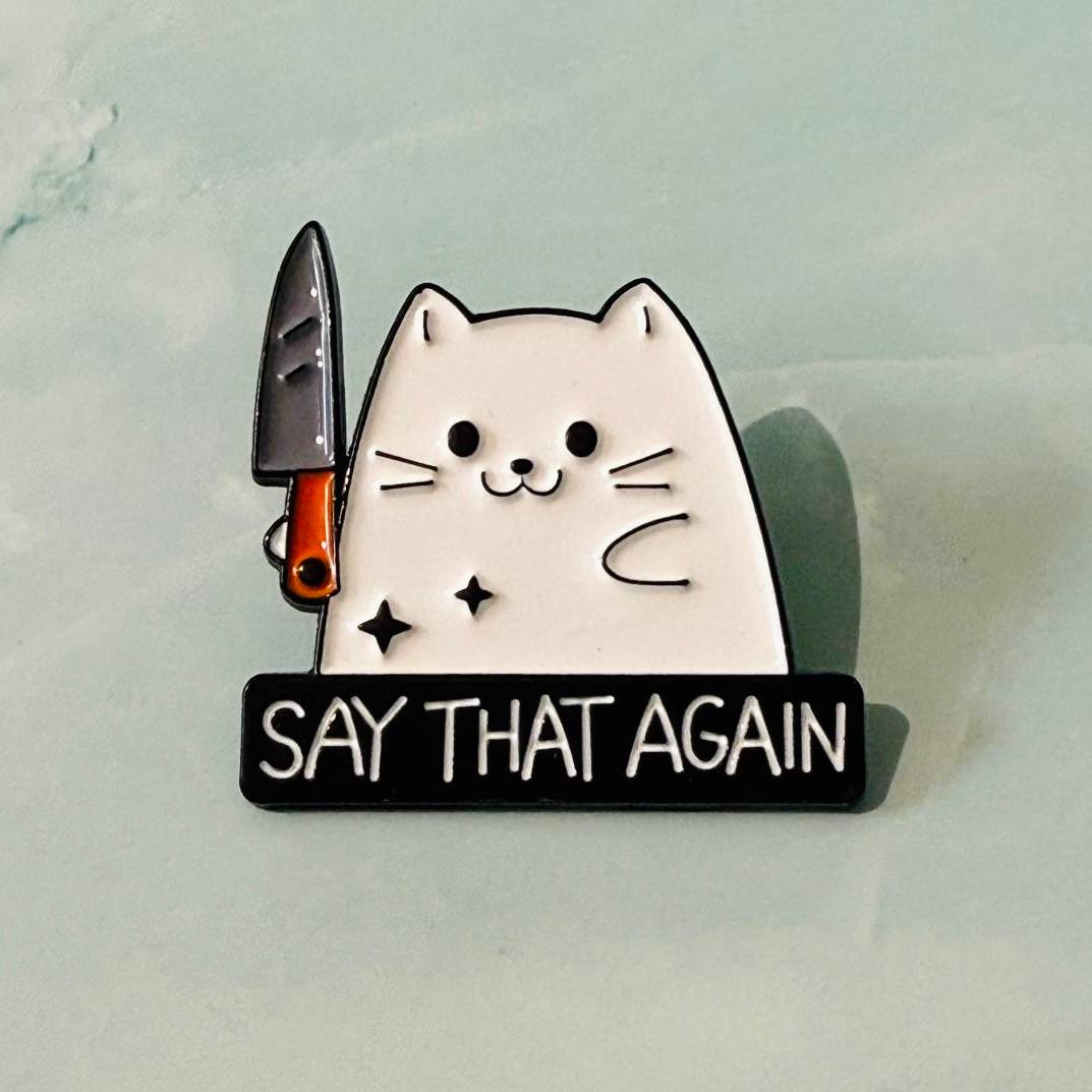 Cat-themed enamel pin with a knife and 'Say That Again' text on a light background