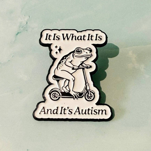 Pin with a frog on a scooter and text 'It Is What It Is And It's Autism' on a marble background